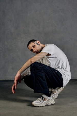 Flexible man with tattooed body and face, earrings, beard. Dressed in white t-shirt and sneakers, black sports trousers. Squatting sideways against gray background. Dancehall, hip-hop. Close up