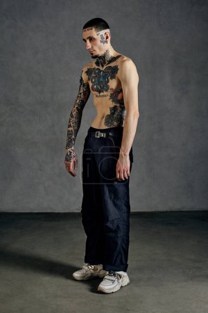 Strong performer with tattooed body and face, naked torso, beard. Dressed in black pants and white sneakers. He is dancing against gray studio background. Dancehall, hip-hop. Full length, copy space