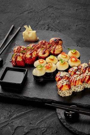 Set of delicious baked uramaki and norimaki topped with golden brown cheese hats traditionally served with pickled ginger and soy sauce on black slate boards. Japanese cuisine