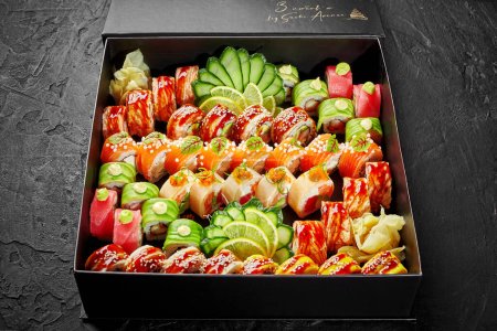 Großes Set verschiedener japanischer Rollen mit Thunfisch, Lachs und Aal, verpackt in Karton mit eingelegtem Ingwer, Gurkenscheiben und Zitrone, auf schwarzem Hintergrund. Sushi-Lieferkonzept