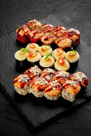 Appetitlich gebackene Sushi-Rollen mit cremigen Käsehüten, gewürzt mit würzigem Mayo und Unagi-Sauce, garniert mit Parmesan-Krümeln, Gemüsespänen und roten Chilifäden auf Schiefertafel