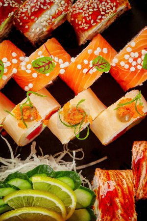 Top view of vibrant assorted sushi rolls with salmon, eel, tuna, mamenori garnished with masago, sesame and airy rice balls accompanied by shredded daikon, cucumber and lime slices on black background