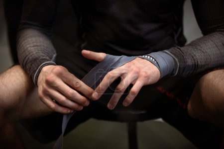 Male boxer sitting in dimly lit gym wrapping hands with protective boxing handwraps before training session to ensure safety and support, cropped image