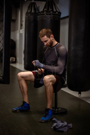 Concentrated young adult bearded fighter in sportswear sitting in dimly gym among hanging boxing bags, meticulously wrapping bandages on wrists, preparing for training fight