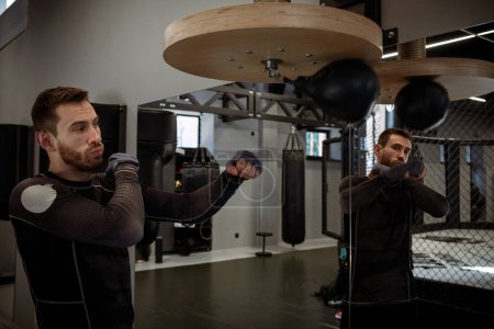 Photo for Concentrated young adult bearded boxer honing skills on speed bag, working out focus and accuracy in well-equipped boxing gym - Royalty Free Image