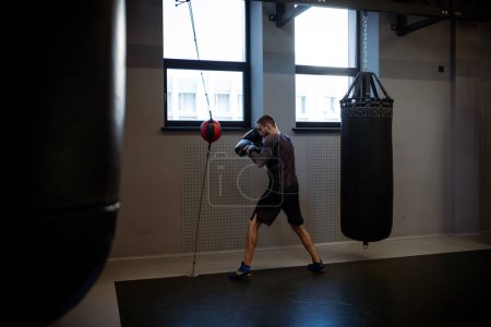 Photo for Determined adult man in sportswear and boxing gloves training with floor to ceiling bag, strengthening muscles, improving reflexes and punching speed in gym setting - Royalty Free Image