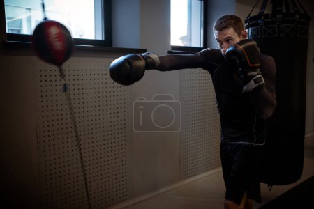 Photo for Motivated young adult mixed martial artist sharpening accuracy and timing using double-end bag at modern boxing gym, demonstrating focus and technique - Royalty Free Image