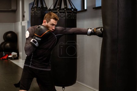 Photo for Young adult boxer focused on improving punch strength and power practicing on heavy bag while training alone in modern boxing gym. Sports motivation concept - Royalty Free Image