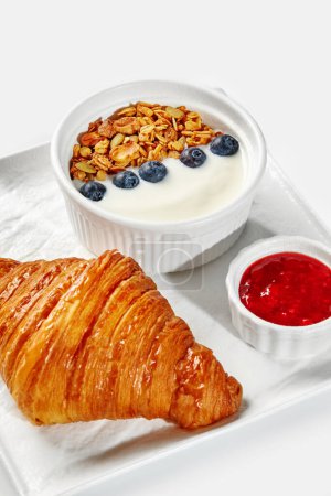 Foto de Delicioso desayuno continental con croissant mantecoso, yogur cubierto con granola y arándanos, y lado de mermelada de bayas rojas servido en bandeja blanca - Imagen libre de derechos