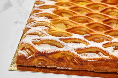 Close-up of golden lattice apple pie with tender custard and powdered sugar, freshly baked and displayed on golden cardboard on white surface