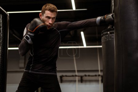 Photo for Motivated young adult mixed martial artist sharpening accuracy and strength using heavy bag at modern boxing gym, demonstrating focus and technique - Royalty Free Image