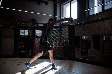 Photo for Male athlete in sportswear and boxing gloves shadowboxing, highlighted by rays of sunlight in well equipped MMA cage in dimly lit training room - Royalty Free Image