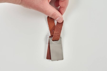 Main masculine présentant un porte-clés en cuir marron non marqué conçu pour l'impression photo ou la gravure personnalisée, sur fond de lumière avec copyspace. Accessoires élégants fabriqués à la main
