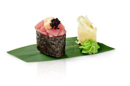 Tuna gunkan sushi topped with caviar and mayo, accompanied by wasabi and pickled ginger, presented on green bamboo leaf against white background. Traditional Japanese cuisine