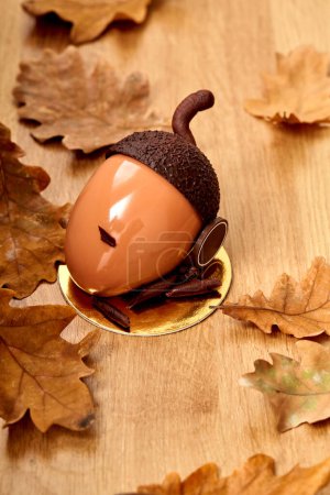 Acorn shaped cake in glossy hazelnut praline ganache and dark chocolate cap and stem, served on golden cardboard on wooden table surrounded by dried oak leaves. Bounties of nature in designer desserts
