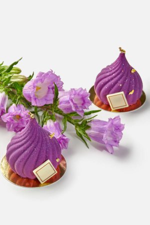 Two purple velvety swirl-shaped pastries decorated with edible gold and chocolate, displayed on gold cardboard cake bases accompanied by sprig of bellflowers on white background
