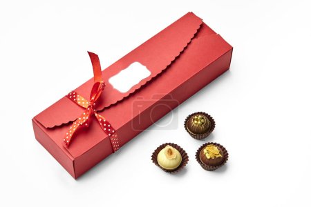 Kunsthandwerkliche Schokoladenbonbons mit Nüssen, Karamellspänen und goldenen Zuckerperlen in eleganter roter Schachtel mit Signatur-Etikett, gebunden mit gepunktetem Band zum Verschenken