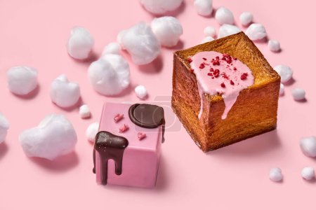 Foto de Dos croissants en forma de cubo con glaseado de baya cremosa suave, adornado con sello de chocolate y trozos de frambuesa liofilizados rodeados de suaves nubes de algodón blanco de azúcar en la superficie rosa - Imagen libre de derechos