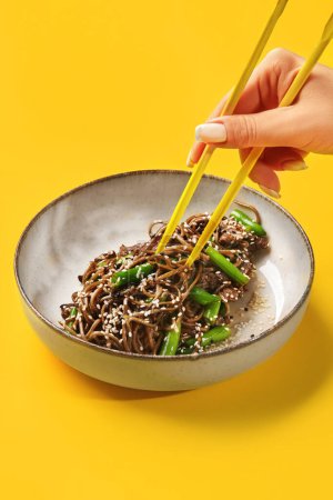 Main de femme utilisant des baguettes pour ramasser des nouilles soba au boeuf frit avec haricots verts et sésame, servies dans un bol en céramique sur fond jaune vif. Plat de cuisine asiatique populaire