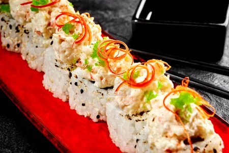 Nahaufnahme von Sushi-Rollen garniert mit Frischkäse-Garnelen-Mischung, garniert mit pikanten Chilispänen und grünem Tobiko-Rogen auf rotem Teller, auf schwarzer Oberfläche mit Essstäbchen und Sojasauce