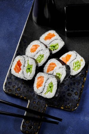 Délicieux rouleaux futomaki au saumon et concombre au design yin-yang traditionnellement servis avec de la sauce soja sur une planche de service contre une surface texturée bleue. Présentation créative des sushis japonais