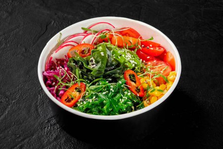 Colorful vegan poke bowl with sliced avocado, radishes, tomatoes, hiyashi wakame, corn, greens and hot chili peppers sprinkled with sesame seeds, presented in cardboard cup on dark textured surface