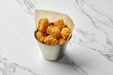 Goldene knusprige Chicken Nuggets in einem mit Pergamentpapier ausgekleideten Eimer aus Edelstahl, präsentiert auf Marmoroberfläche. Beliebtes Fast-Food-Gericht