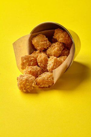 Crispy fried chicken nuggets spilling from parchment-lined metal bucket onto sunny yellow background. Popular food from menu of fast food restaurants