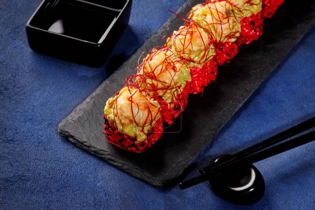 Vibrant red rice sushi rolls with topping of avocado spread and tempura fried shrimp garnished with togarashi threads, elegantly presented on black slate serving board with soy sauce. Japanese cuisine
