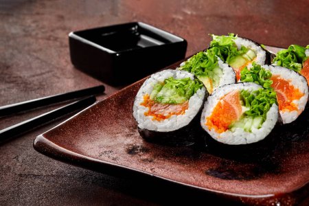 Appetitlich bunte Lachs- und Gurken-Maki-Sushi-Rollen mit Tobiko und Salat, traditionell auf rustikalem Teller mit Sojasauce serviert. Japanische Küche