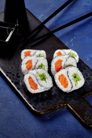 Set of yin and yang shaped futomaki rolls filled with salmon and crisp cucumber served on textured plate with chopsticks and soy sauce against blue background. Japanese style snack