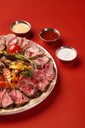 Assorted meat platter with roast beef and baked pork loin garnished with fresh aromatic herbs, served with mix of grilled vegetables and marinated onions, accompanied by dips against red backdrop