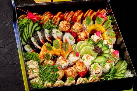Foto de Lujoso juego de rollos de sushi con diversos rellenos y coberturas, ingeniosamente dispuestos en caja de cartón adornada con flores, daikon rallado, pepino y lima sobre fondo oscuro. Concepto de comida para llevar - Imagen libre de derechos
