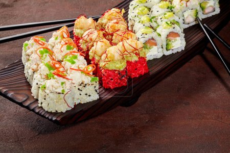 Verlockende Sushi-Rollen mit Lachs, Garnelen, Tobiko und Sesam garniert mit Gemüse und würziger Sauce, arrangiert auf einem dunklen Holztablett. Stilvolle Präsentation beliebter japanischer Gerichte