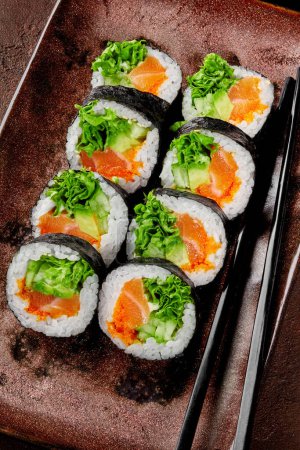 Delicious futomaki rolls with fresh salmon, tobiko, avocado, cucumber and lettuce served on rustic plate with chopsticks. Popular Japanese snack