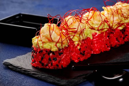 Closeup of red rice sushi rolls coated with black sesame, topped with avocado spread and deep-fried shrimp and garnished with togarashi threads, served on black slate board with soy sauce