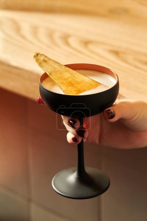 Female hand elegantly holding black stemmed glass with sweetish light alcoholic cocktail garnished with spicy caramelized pear slice against blurred background of wooden bar counter