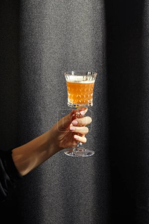 Mano femenina elegantemente sosteniendo cristal lleno de whisky agrio, cóctel en tono ámbar en contraste con el fondo de tela de textura oscura