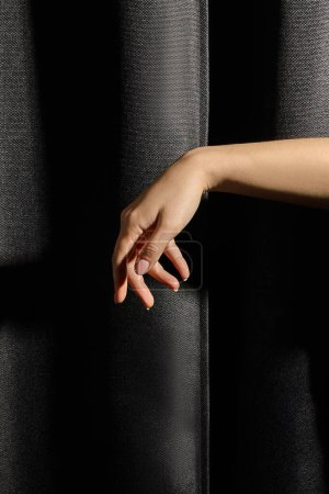 Graceful female hand in soft natural pose contrasting with texture of black curtain in dramatic lighting. Minimalist scene evoking themes of elegance, solitude, subtle interplay of light and darkness