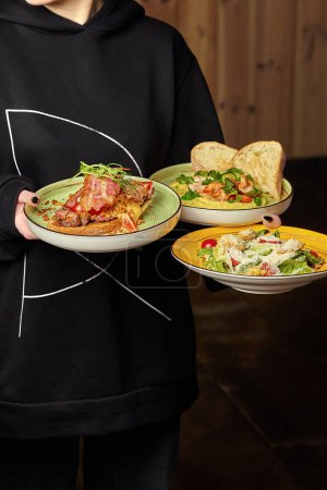 Waitress in black hoodie expertly balancing three plates of appetizing gourmet bruschetta with veal steak and bacon, creamy hummus with shrimp and greens, and classic Caesar salad, cropped shot