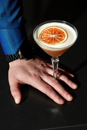 Élégants messieurs main appuyée contre la surface noire, tenant un verre de cocktail aigre-amande avec un haut mousseux orné de tranches de pamplemousse séchées