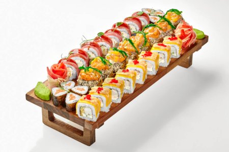Wooden serving tray with various sushi rolls with salmon, tuna, cheese garnished tobiko, hiyashi wakame and greens accompanied by pickled ginger and wasabi, presented isolated on white background