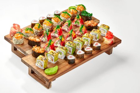 Verlockende Auswahl an verschiedenen japanischen Sushi-Rollen und Gunkan Maki mit Avocado, Tobiko, Lachs, Thunfisch und Hiyashi Wakame, traditionell serviert mit würzigem Wasabi und eingelegtem Ingwer auf Holztablett auf weißem
