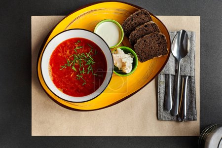 Schüssel mit klassischem Borschtsch mit Kalbfleisch und Bohnen garniert mit Gemüse, serviert mit Roggenbrot, saurer Sahne und Knoblauchschmalzaufstrich, auf gelbem Ladeteller und Papier-Tischset, von oben. Ukrainische Küche