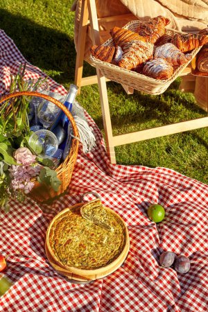 Appetizing savory quiche and sweet puff croissants with fresh fruits and bottle of wine arranged on wooden chair and red and white checkered blanket on lush green grass during summer picnic.