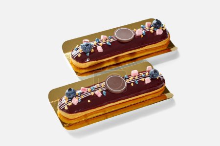 Photo for Two delicious chocolate glazed eclairs topped with fresh blueberries and decorative sweets on gold serving cardboard. Popular French pastry - Royalty Free Image