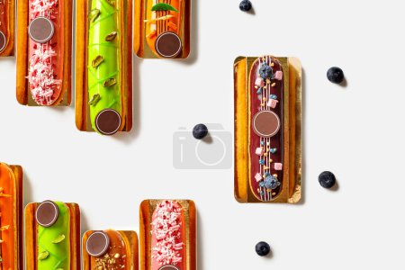 Assortiment de tentantes éclairs français décorés avec des glaçures colorées vibrantes surmontées de baies fraîches, de fruits secs, de noix et de morceaux de meringue, disposés sur des cartons de service dorés sur blanc, vue sur le dessus