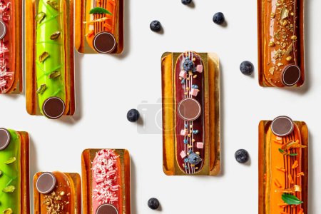 Delicious eclairs with colorful icing and various toppings garnished with fresh blueberries, citrus and chocolate shavings, nuts and decorative elements, on white surface. French style sweets