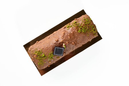 Top view of delicious artisanal loaf cake coated with milk chocolate and nuts glaze, decorated with pistachio crumble and dark chocolate branding plaque, isolated on white. Sweet pastries concept