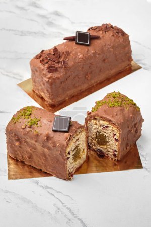 Sliced artisanal loaf cake with fruit jam filling and dried berries, coated with milk chocolate and nuts glaze, decorated with dark chocolate branding plaque and pistachio crumble on golden cardboard